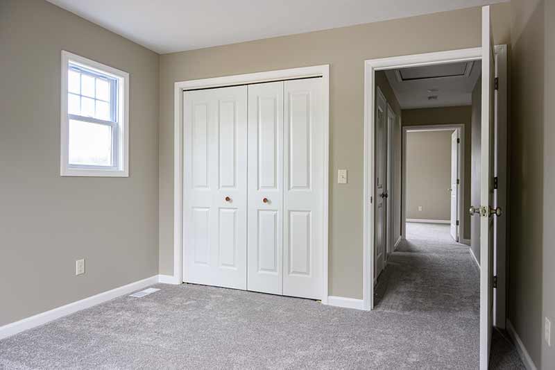 The interior of a carpeted bedroom with a closet and small window. To the right of the closet is a opened door that looks down a short hallway to another bedroom.