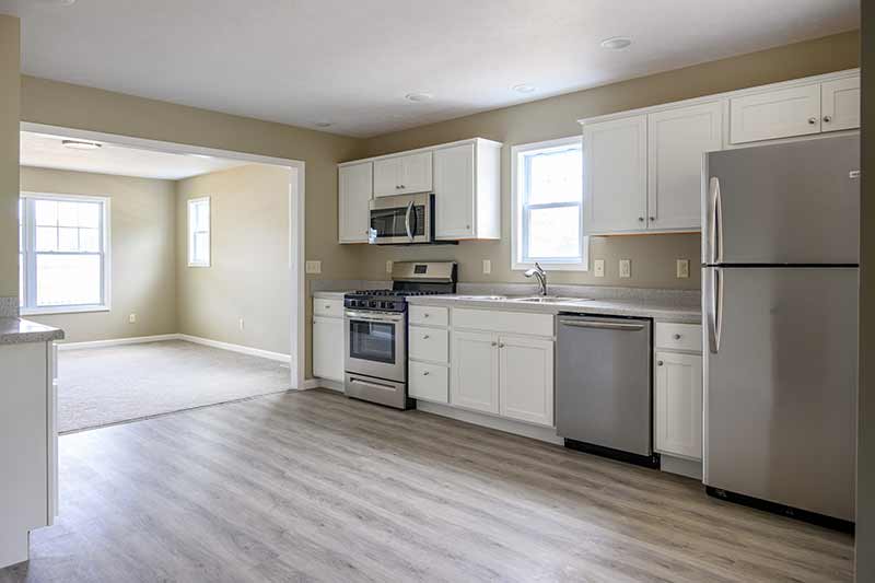 The interior of a newly built kitchen. White cabinates line the far wall along with a fridge, dish washer, sink, oven, and microwave. An empty carpeted living room can be seen towards the back.