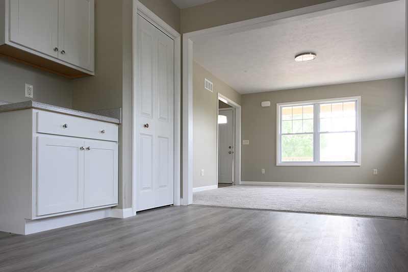The interior of a section of the kitchen laminate wood floor flows into the carpeted living room. In the living room next to the front door is a large window with light shining in.