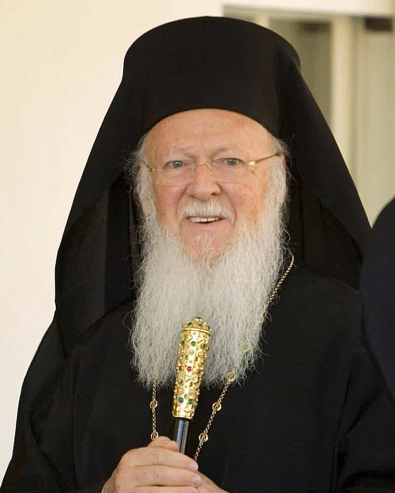 Patriarch Bartholomew I wearing black religious attire, including a headpiece and gold necklace. He's holding a gold object, wears glasses and has a long white beard.