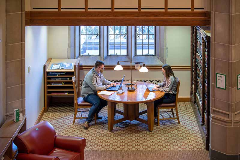 Two masked students on laptops work at a table together.