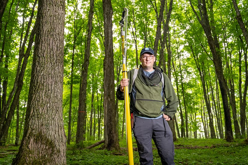 Nate Swenson standing with a pole clipper in a thick wooded forest with mosquitoes swarming around and on him.