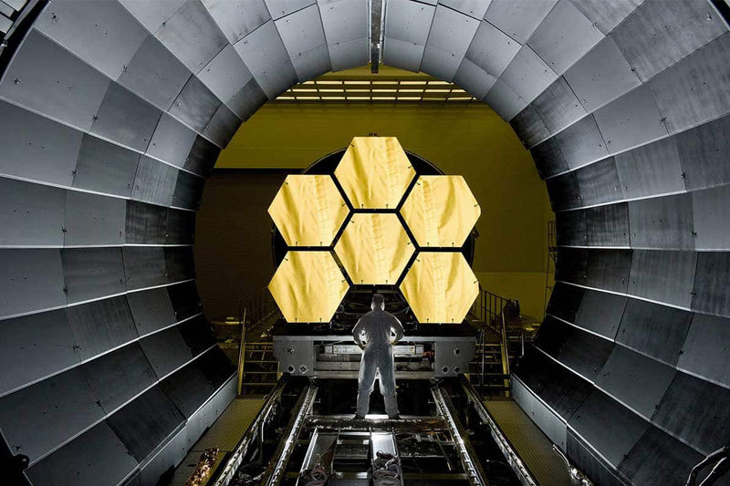 A person with their back to the camera stands in front of a part of Webb's telescope featuring 6 golden hexagons, wearing a cleanroom suit stands with their legs shoulder-length apart, their hands on their hips.