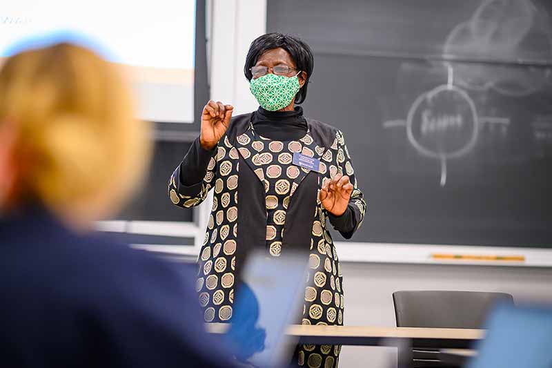 A woman wearing a face mask presents in a classroom in front of a chalkboard, with a student in the foreground.