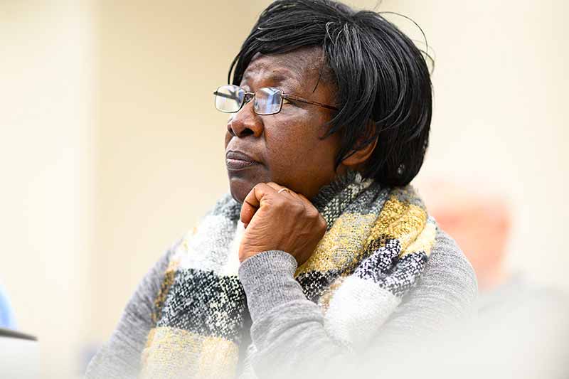 A woman wearing glasses and scarf listens in class with her hand tucked under her chin.