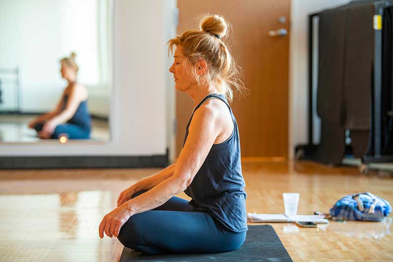 A woman sits on a yoga mat with her hands resting on her crossed legs.