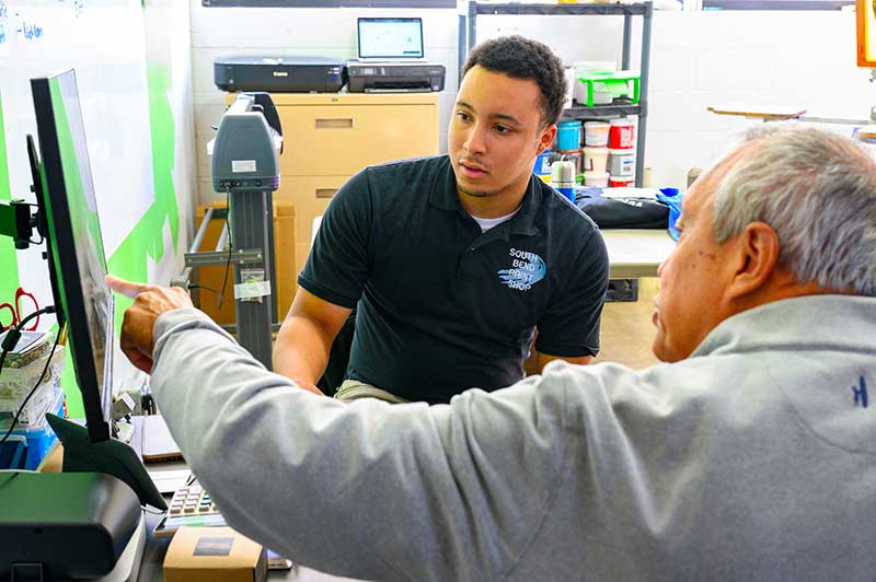 An older man points to a computer screen, showing the younger man something.