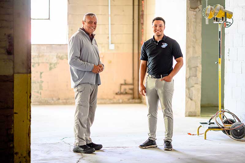 An older man stands next to a younger man, wearing a company shirt, in a warehouse.