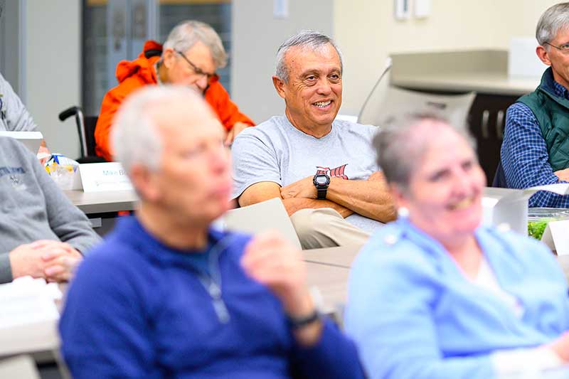 An older man smiling in a classroom with his arms crossed.