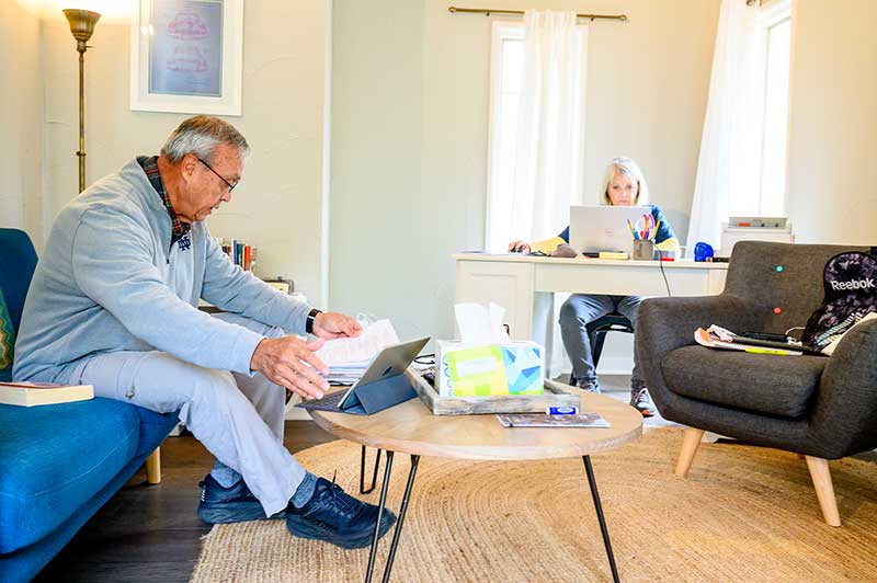 A man reads at a coffee table on the couch, and a woman works at a laptop at a table in the background.