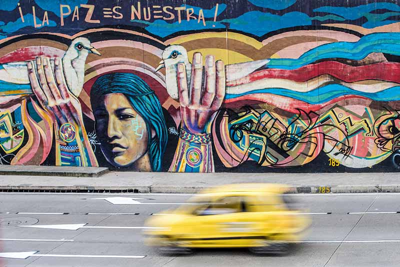 Colorful graffiti on a wall next to a highway. A woman is holding up two doves and a saying above her says 'Peace is Ours'.