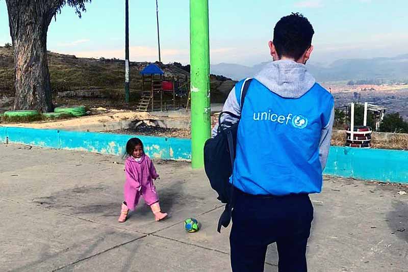 Juan wearing a bright blue UNICEF jacket, watches a child play with a soccer ball.