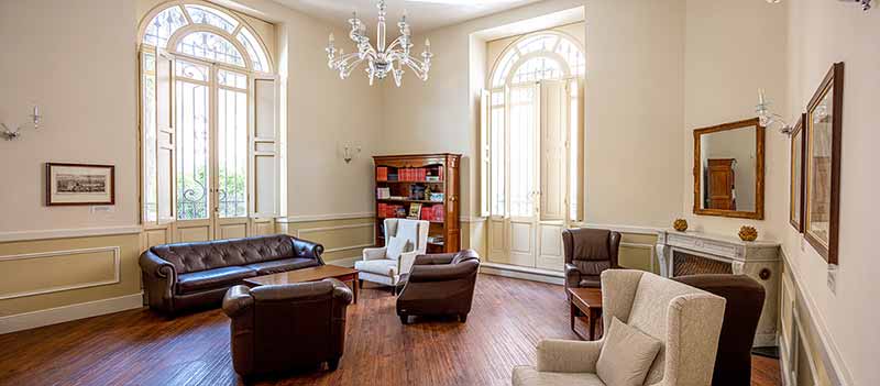 A bright and beautiful study room with leather couches and chairs. A chandelier hangs from the ceiling and open windows bring in the light.