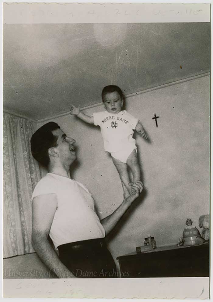 Apartment 22C bedroom - With one hand, George Barbero holds up his son Emmett (3 months), wearing a Notre Dame tee-shirt, August 1952.