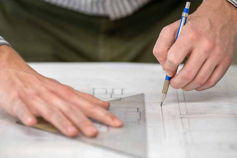 A close up of someone creating a architectual sketch. One hand holds onto a triangular ruler and the other draws a straight line with a pencil.