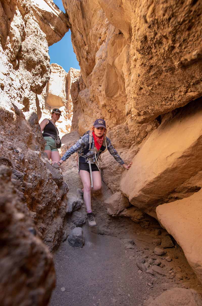 Two female students climb down a narrow passage between yellow rock formations.