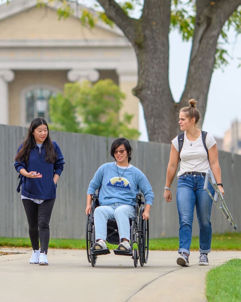 A student using a wheelchair rides down the sidewalk between two people, one who is carrying crutches.