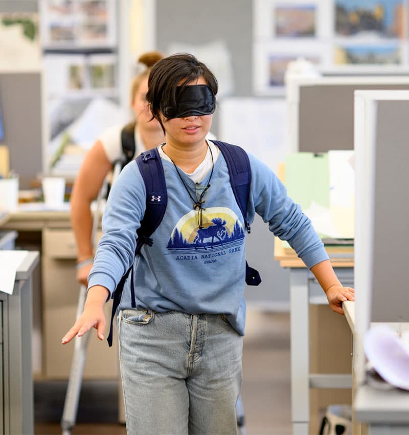 A student wearing a blindfol attempts to move through rows of desks in a clasroom.