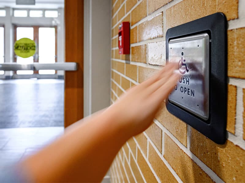 A hand presses a handicap button to open a door.