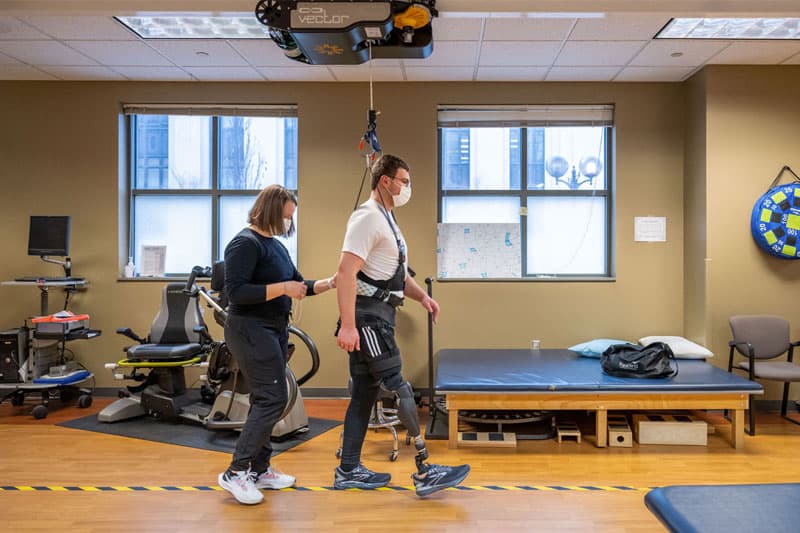 A man is strapped into a harness that is suspended by the ceiling, helping him learn to walk with the prosthetic.