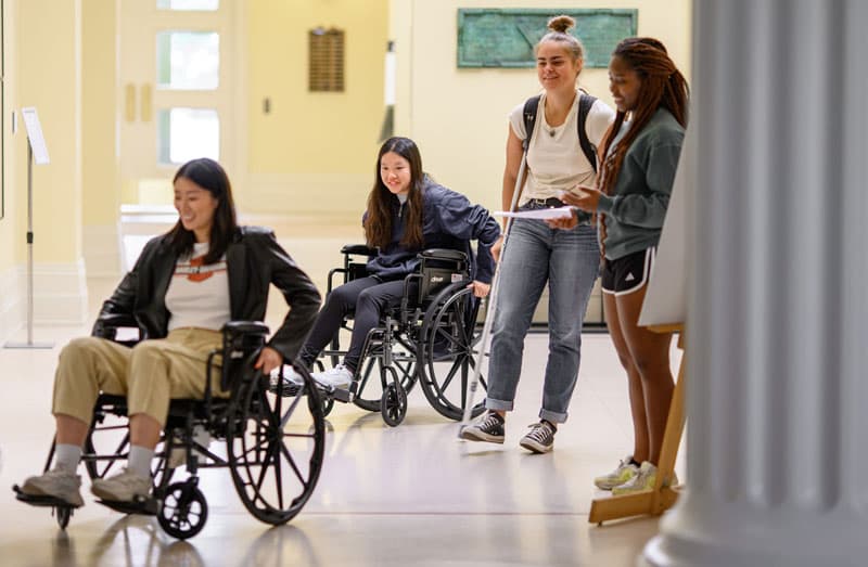 Two students in wheelchairs and a student using crutches move through the hallway in Walsh hall.
