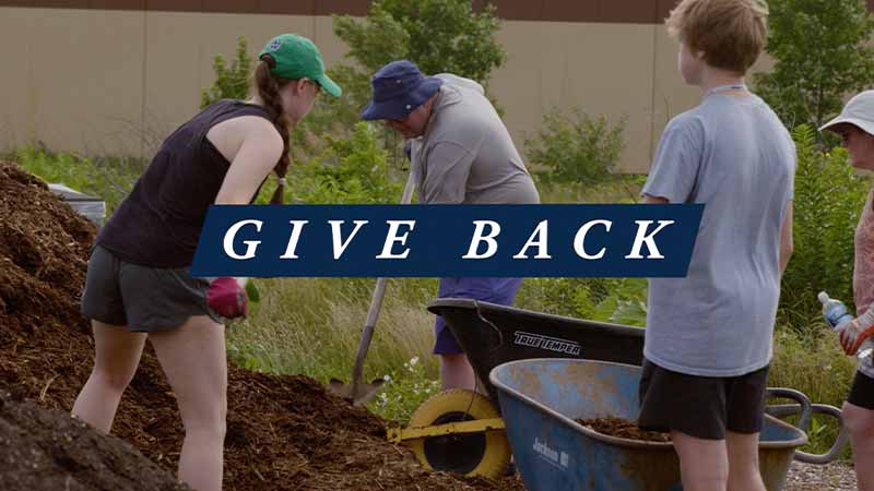 A small group of people use shoves to move dirt to a wheelbarrel. 'Give Back' text on top of the image.