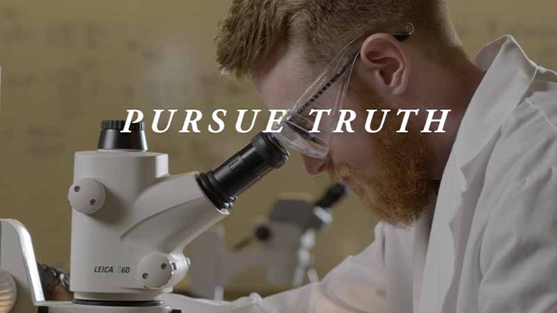 A White man with red hair wearing glasses and a white lab coat looks into a microscope. 'Pursue Truth' text on top of the image