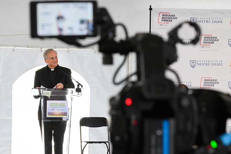 University of Notre Dame President speaks at a podium focused in the background, a camera out of focus stands in the foreground.