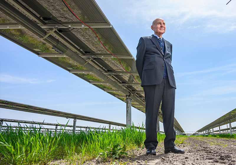 An elderly man stands next to solar panels looking off camera.