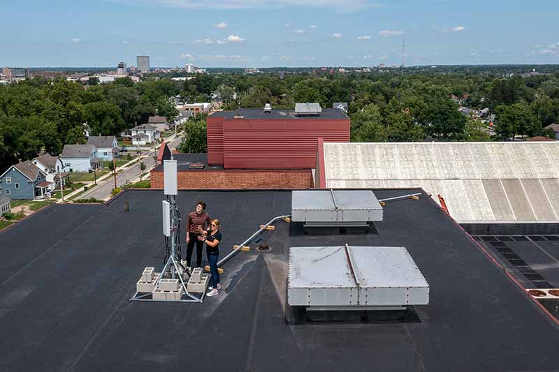 A man and a woman stand on top of a school roof and inspect an antenna used to create a wifi network. Residental homes can be seen in the background.