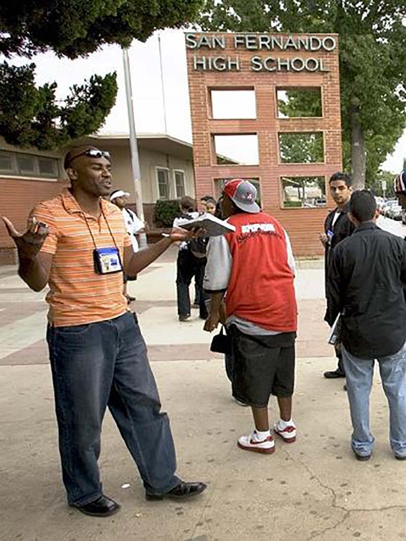 Morrell holds a book and gestures to students outside of a brick building signed San Fernando High School.