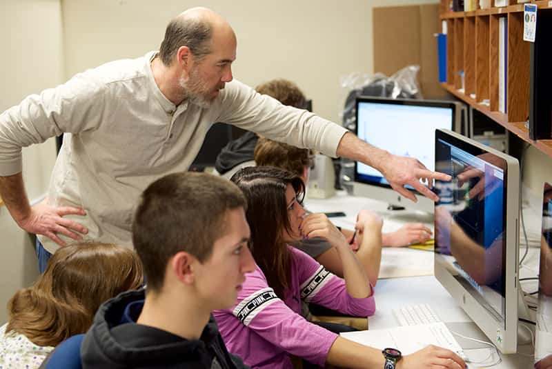 A male teacher points to a computer, helping a student.