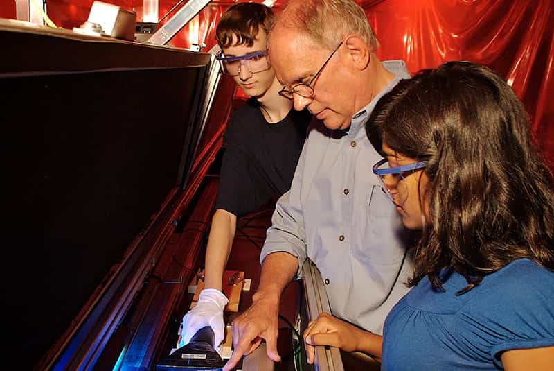 A teacher and two students wearing eye googles examine a device.