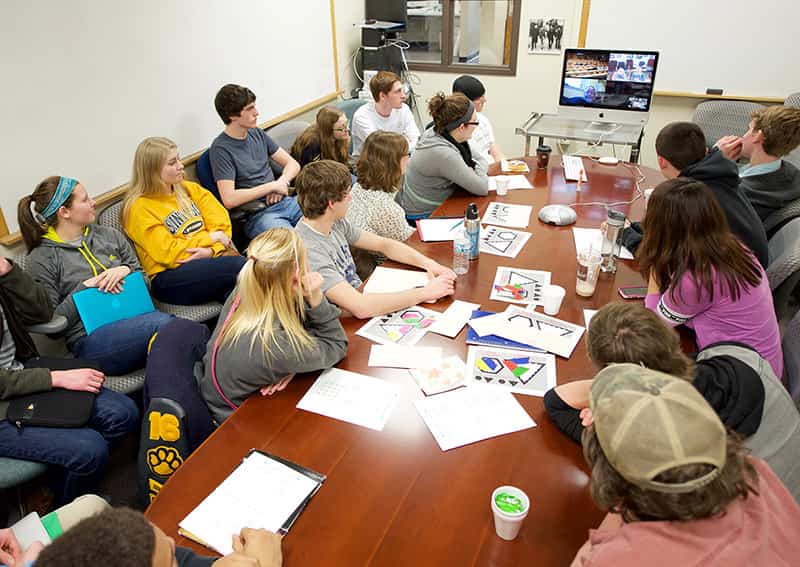 Students gather around a large, rectangular table with a computer sitting at the front and four other people on screen for a video conference.