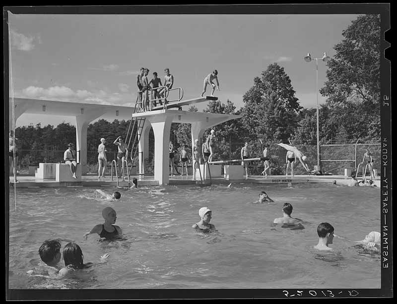 An archival photo of only white people swimming and diving off of a diving board at a community swimming pool.