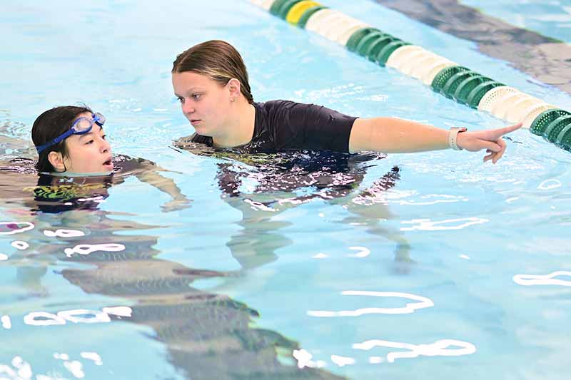 A student athlete points forward while talking with a middle schooler in a pool.
