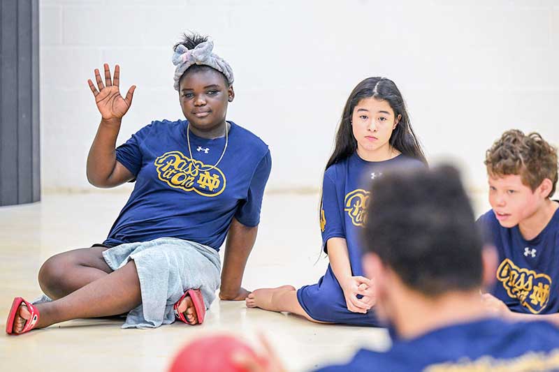 Three middle schoolers sit on the ground. One raises her hand.