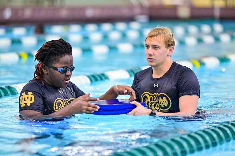 A student athlete and middle schooler stand next to each other in a pool holding onto a kickboard.