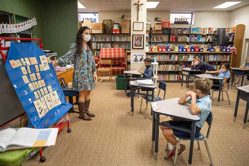 A masked teacher stands in front of a class of masked students.