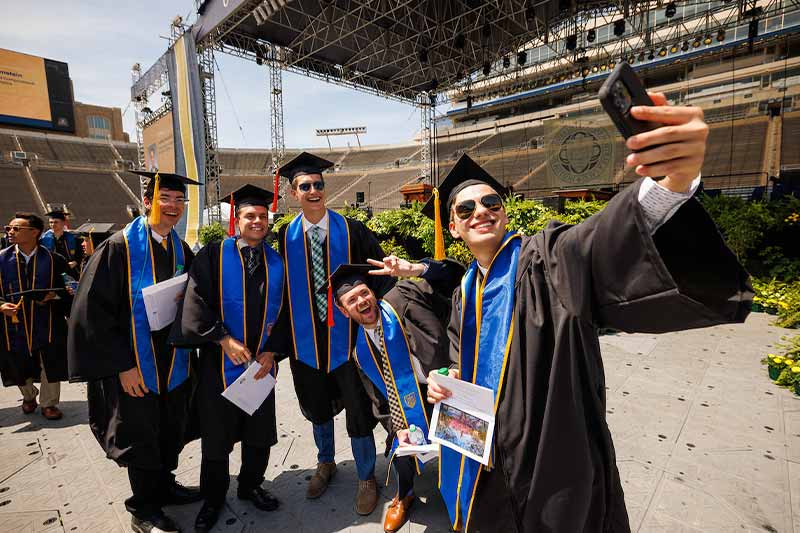 Five men wearing caps and gowns take a selfie in the Notre Dame stadium.