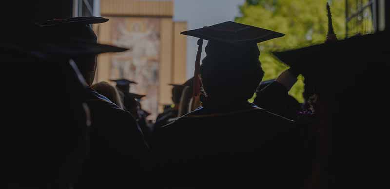 A silhouette from behind of graduates wearing caps and gowns looking towards Touch Down Jesus.