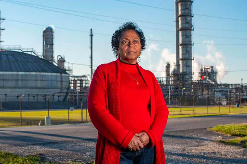 Sharon Lavigne, a black woman wears a red sweater and cross necklace stands in front of a chemical plant looking off camera.