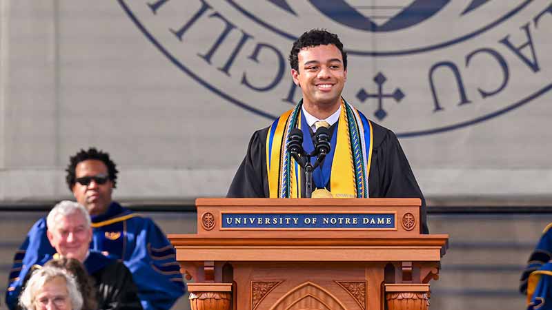 Devin Diggs, a man wearing a black graduation robe with a number of academic cords and stoles in blue and gold, speaks at a podium.