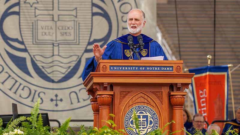 Archbishop Borys Gudziak wears navy blue commencement regalia of the University of Notre Dame as he speaks at a podium at a commencement ceremony. Others wearing the same robes sit behind him.