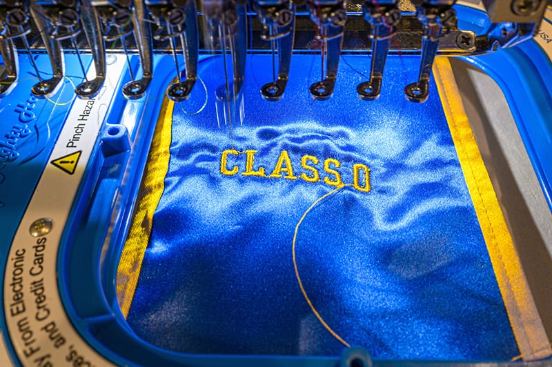 An embroidery machine stitching 'Class of 2023' on a blue and gold stole.