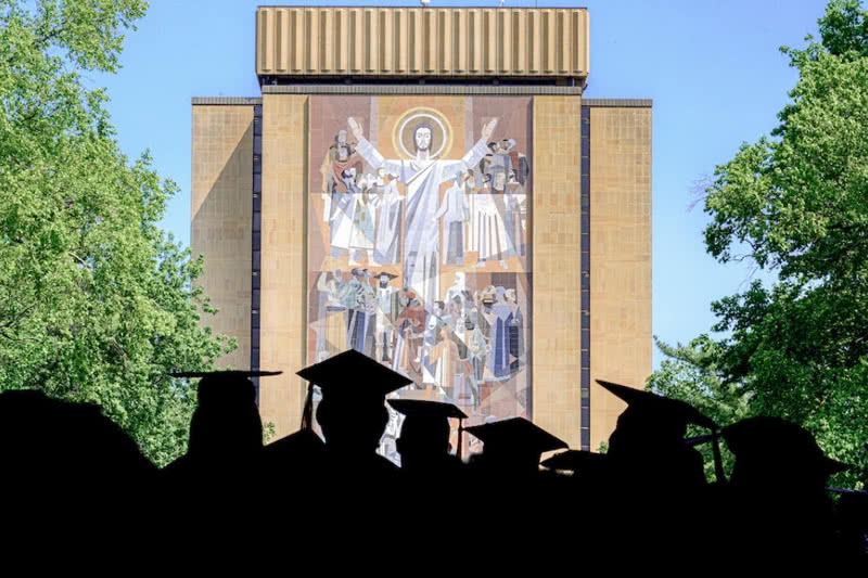 Silhouettes of graduates in caps and gowns with the Hesburgh Library in the background.