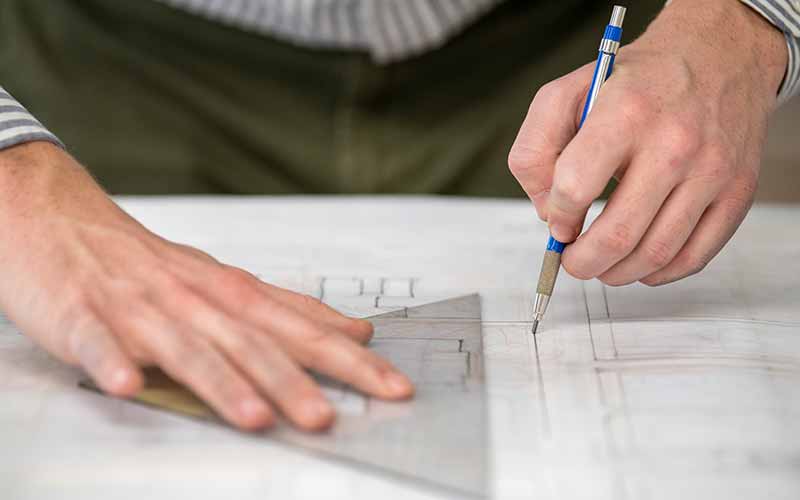 A close up of someone creating a architectual sketch. One hand holds onto a triangular ruler and the other draws a straight line with a pencil.