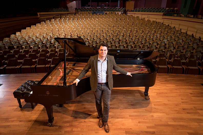 J.J. Wright standing in front of a piano, on stage in a concert hall