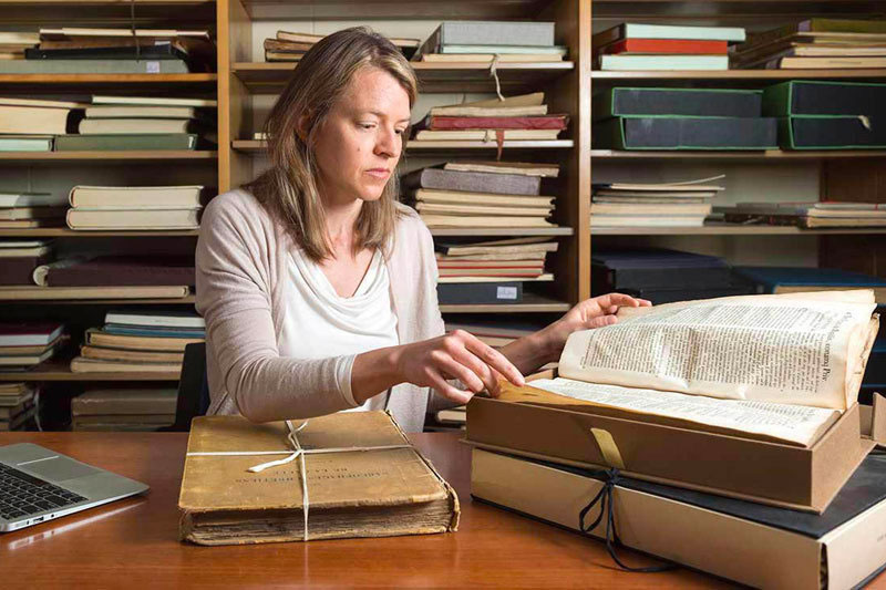 Elaine Stratton Hild sits at a table in front of bookshelves while looking at ancient textbooks.