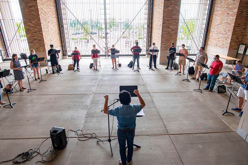 A group of choir students stand inside the Leahy Gate of the football stadium with music stands in front of them.